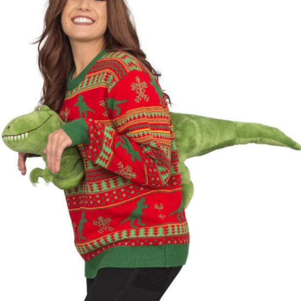 Eine Frau trägt einen roten Weihnachtspullover mit Dinosaurier-Motiv und hält ein grünes Dinosaurier-Stofftier.