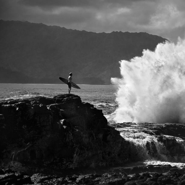 Eine Welle schlägt am Felsen auf. Dahinter steht ein Mann mit Surfbrett unterm Arm.