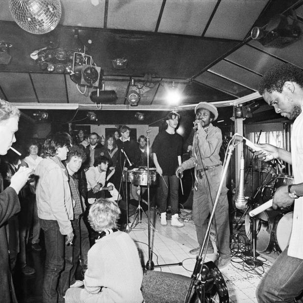 Auf einer kleinen Bühne in einer niedrigen Disco spielt eine Band, da Publikum raucht, sitzt und steht herum.