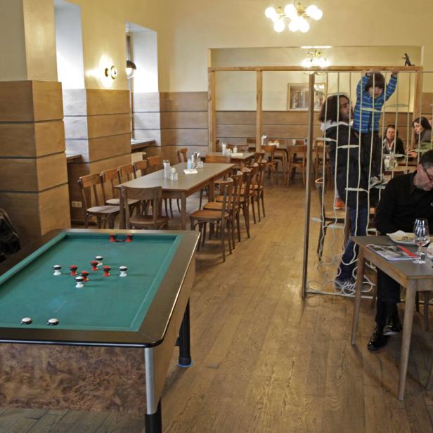 In einem Restaurant spielen Kinder an einem Gitter, während Erwachsene an Tischen sitzen.