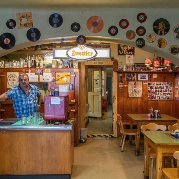 Ein Mann steht hinter der Bar in einem traditionellen Wiener Beisl, dekoriert mit Schallplatten und Bierwerbung.