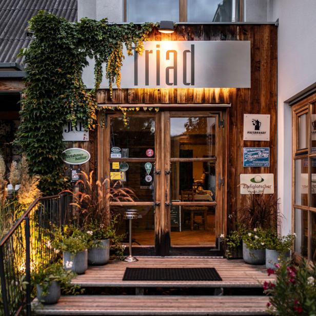 Der Eingang zum Restaurant „Triad“ mit Holzverkleidung und Pflanzen wirkt einladend.