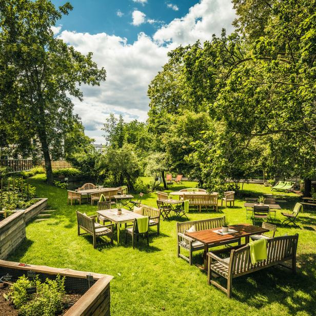Ein Garten mit Tischen und Stühlen unter Bäumen an einem sonnigen Tag.