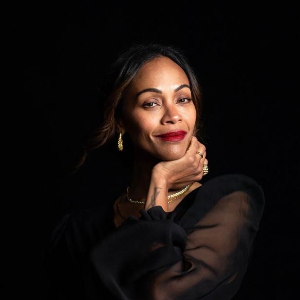 Zoe Saldana posiert vor einem dunklen Hintergrund in einem schwarzen, transparenten Oberteil.