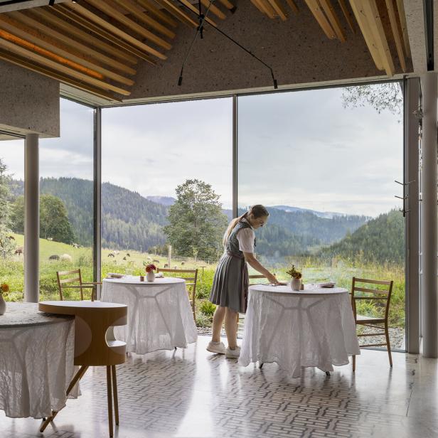 Eine Frau im Dirndl deckt Tische in einem Restaurant mit Blick auf eine grüne Landschaft.