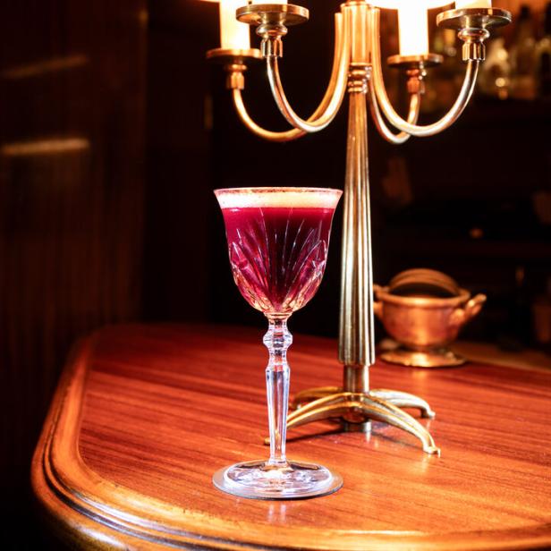 Ein roter Cocktail in einem Kristallglas steht auf einem Holztisch neben einer goldenen Lampe.