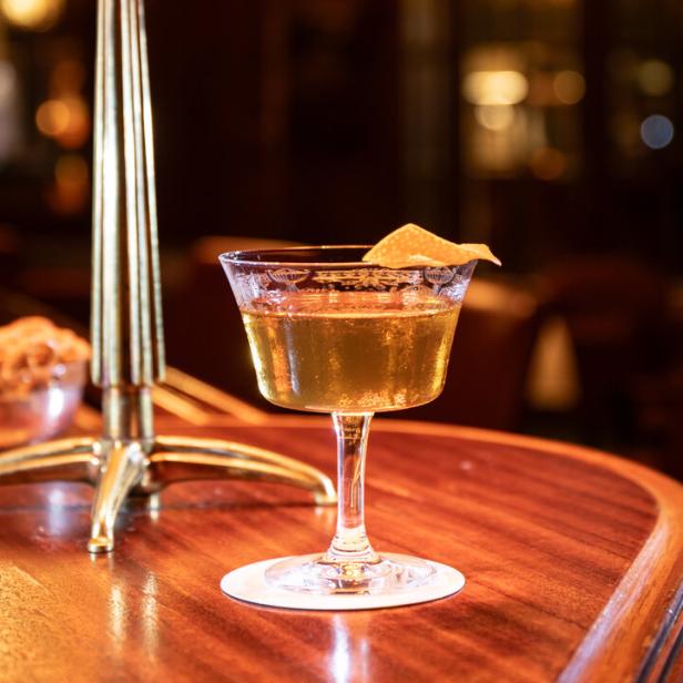 Ein Cocktail mit Orangenzeste steht auf einem Holztisch neben einem Kerzenständer.