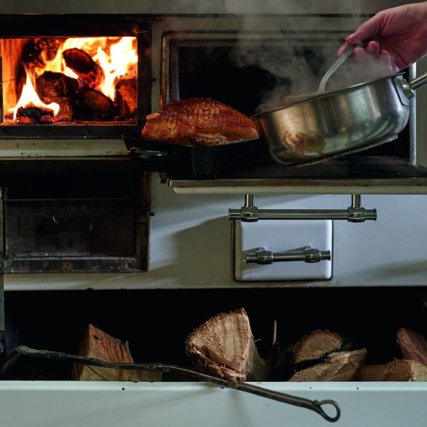 Ein Braten wird in einem Holzofen zubereitet, während eine Person ihn mit Flüssigkeit übergießt.