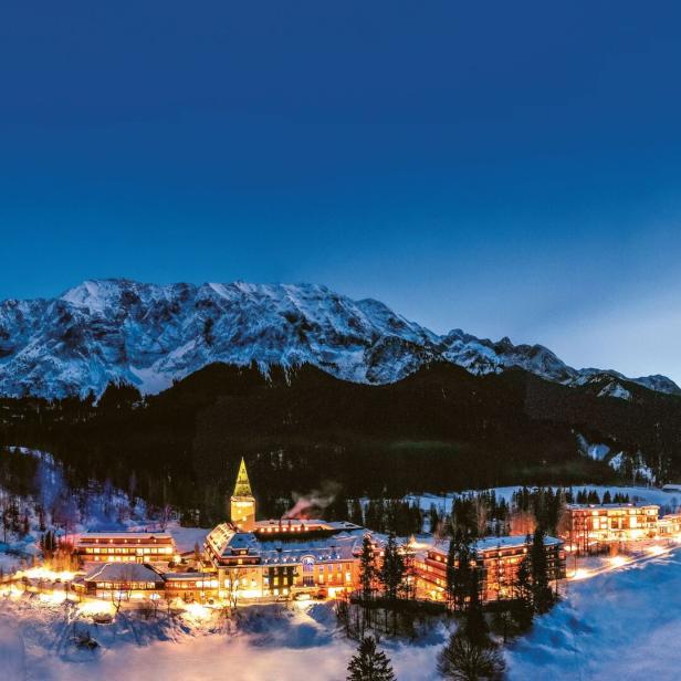 Das beleuchtete Hotel Schloss Elmau im Winter vor einer Bergkulisse.