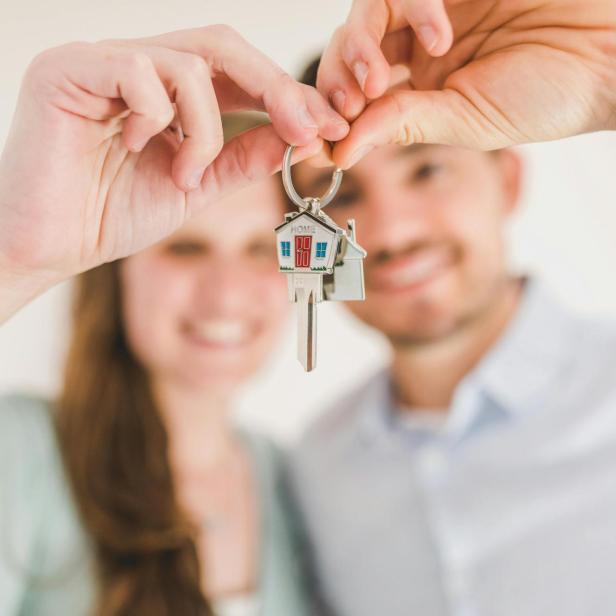 Ein hetero-Paar hält lächelnd einen Schlüssel in die Kamera, der oben geformt ist wie ein Haus.