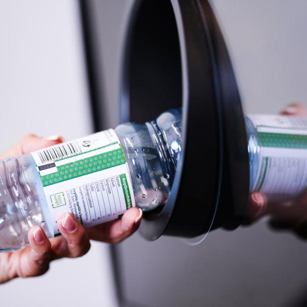 Eine Hand wirft eine leere Plastikwasserflasche in einen Recyclingbehälter.