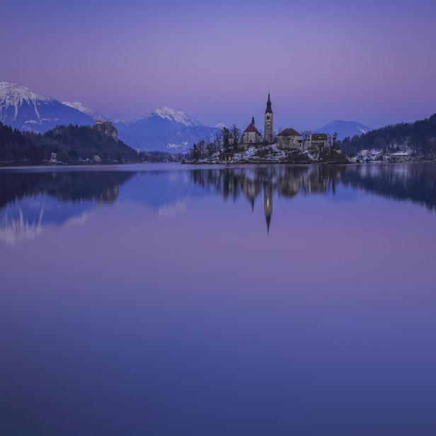 Eine Kircheninsel inmitten des Bleder Sees, aufgenommen am Abend. Rundherum stehen Hügeln und Berge.