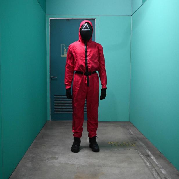 Eine Person in rotem Overall und Maske mit Dreieck steht in einem grünen Gang.