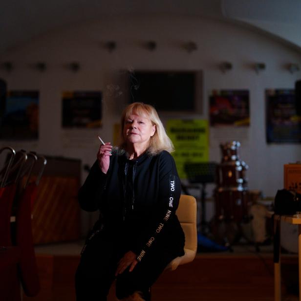 Eine Frau mit blonder Bob-Frisur raucht eine Zigarette in einem Raum mit Musikinstrumenten.