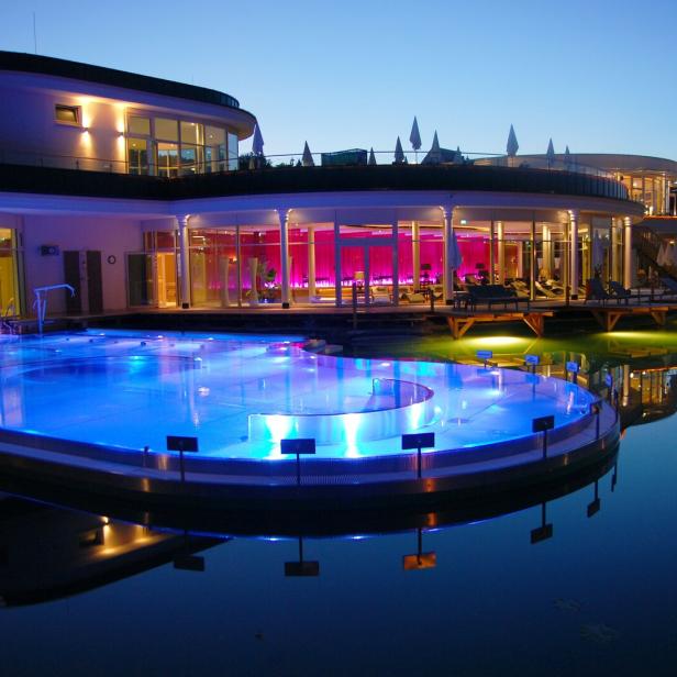 Modernes Wellnessgebäude mit beleuchtetem Außenpool und großen Glasfronten in der Abenddämmerung.