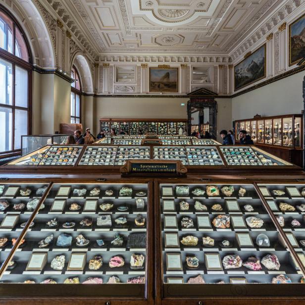 Ein Ausstellungssaal mit großen Vitrinen der Edelstein- und Mineraliensammlung in Wien. An den Wänden hängen große Wandgemälde und Stuck.