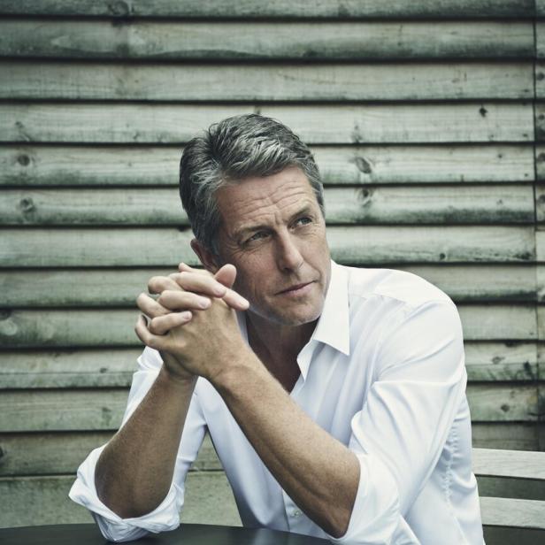 Hugh Grant sitzt an einem Tisch und blickt nachdenklich zur Seite.
