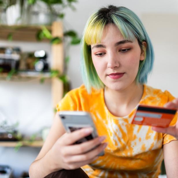 Jugendliche mit kurzen blauen Haaren schaut auf ihr Smartphone und hält eine Kreditkarte in der Hand