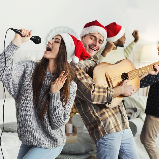 Eine Frau singt in ein Mikrofon, während ein Mann Gitarre spielt und andere im Hintergrund tanzen.