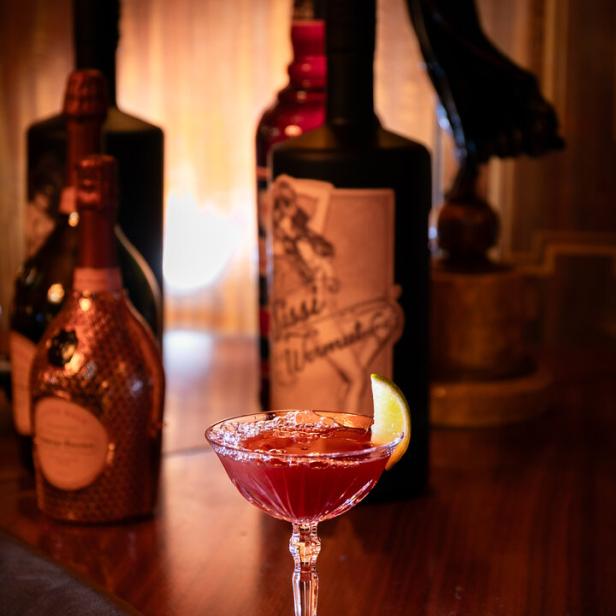Ein roter Cocktail mit Limettenscheibe steht auf einer Bar, im Hintergrund diverse Flaschen.