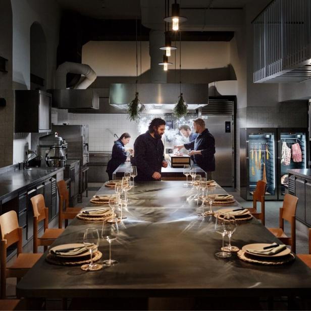 Das Restaurant „Mama Konstantina“ mit Köchen bei der Arbeit und einem langen, gedeckten Tisch.