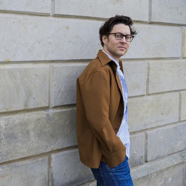 Ein Mann mit Brille und brauner Jacke steht vor einer Steinwand.