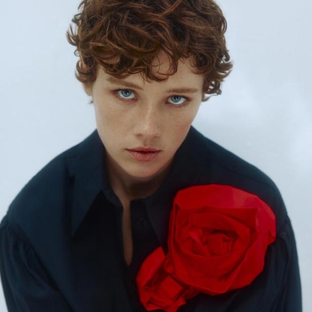 Eine Frau mit roten, lockigen Haaren trägt eine schwarze Bluse mit einer großen roten Stoffblume.