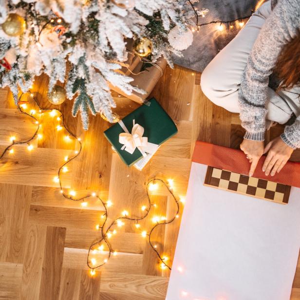 Eine Frau sitzt neben einem Weihnachtsbaum und verpackt ein Schachbrett als Geschenk.