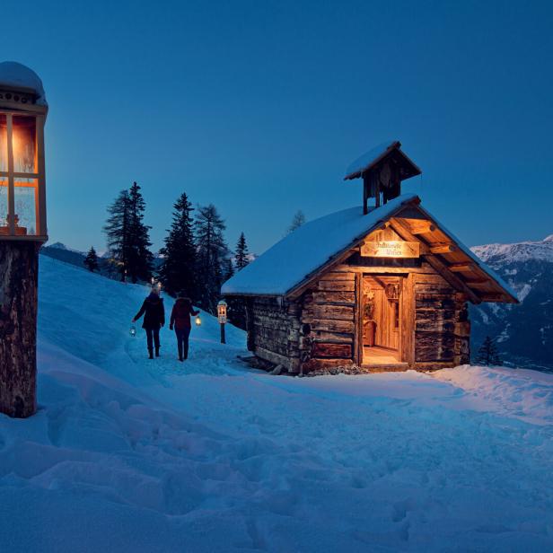 Zwei Personen wandern im Schnee zu einer beleuchteten Holzhütte.