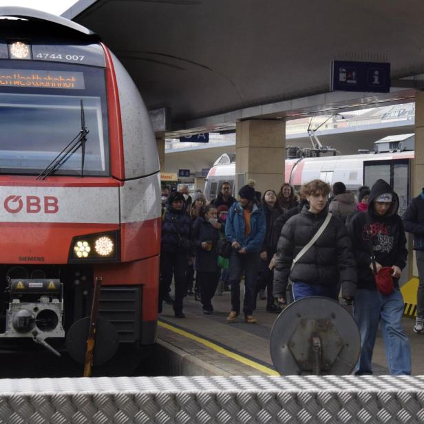 Ein roter Zug steht am Bahnsteig, viele Menschen steigen aus und gehen mit Gepäck und Masken entlang des Bahnsteigs.