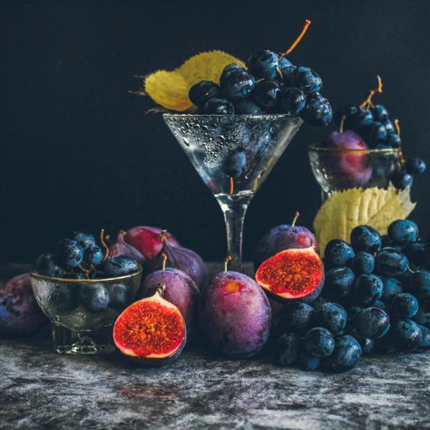 Verschiedene dunkle Trauben, Pflaumen und aufgeschnittene Feigen liegen zusammen mit Blättern auf einer Steinplatte.