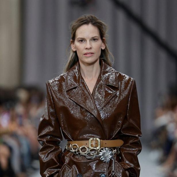 Hilary Swank trägt einen glänzenden, braunen Trenchcoat mit Gürtel auf einem Laufsteg.