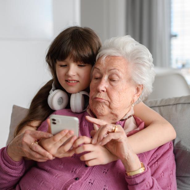 Ein Mädchen mit Kopfhörern umarmt eine ältere Frau, während beide gemeinsam auf ein Smartphone schauen.