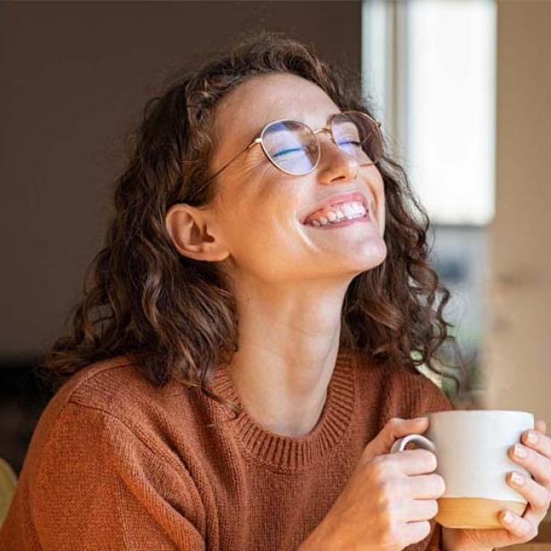 Eine Frau mit lockigen Haaren lächelt der Morgensonne entgegen, während sie eine Tasse Kaffee in der Hand hält. 