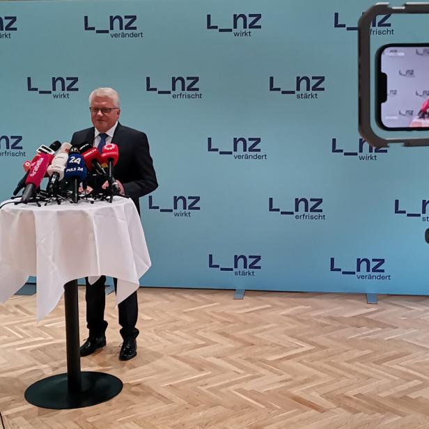 Ein Mann steht vor Mikrofonen bei einer Pressekonferenz, die per Smartphone gefilmt wird.