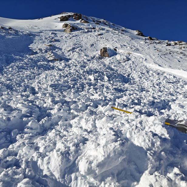 Große Schneemassen und Geröll nach einem Lawinenabgang an einem verschneiten Berghang mit Ausrüstungsgegenständen im Vordergrund.