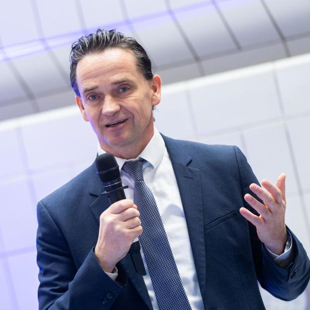 Ein Mann im Anzug hält ein Mikrofon und gestikuliert während einer Präsentation.