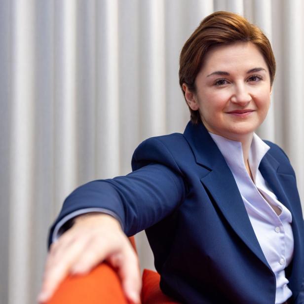 Eine Frau mit kurzem braunem Haar trägt einen blauen Blazer und sitzt entspannt auf einem orangefarbenen Stuhl vor hellen Vorhängen.