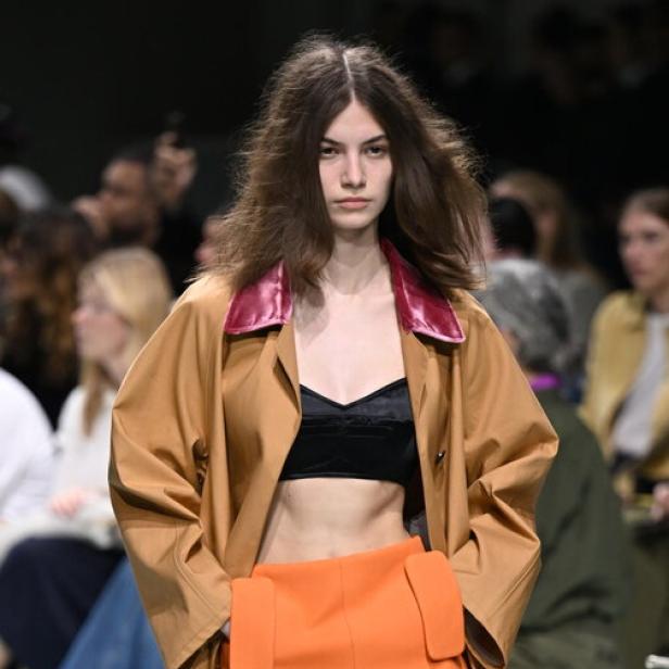 Eine Frau auf dem Laufsteg mit einem Trenchcoat, einem schwarzen Top und einem orangefarbenen Rock.