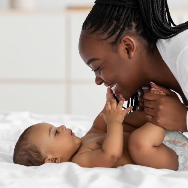 Eine lächelnde Mutter spielt mit ihrem Baby auf einem weißen Bett.