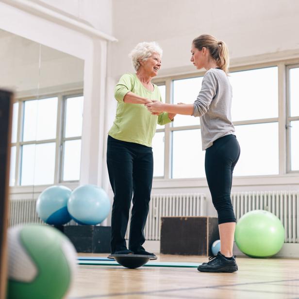 Eine Trainerin hilft einer Seniorin auf dem Balance-Board.