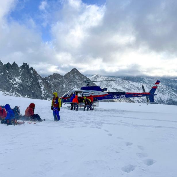 Mehrere Menschen mit Ausrüstung stehen und sitzen im Schnee neben einem Hubschrauber in einer verschneiten Berglandschaft.