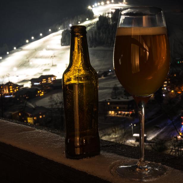 Eine Bierflasche und ein gefülltes Glas stehen auf einer verschneiten Brüstung mit beleuchteter Skipiste im Hintergrund.