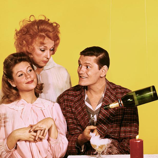 Drei Personen sitzen an einem Tisch, während eine Flasche Sekt in ein rauchendes Glas gegossen wird.