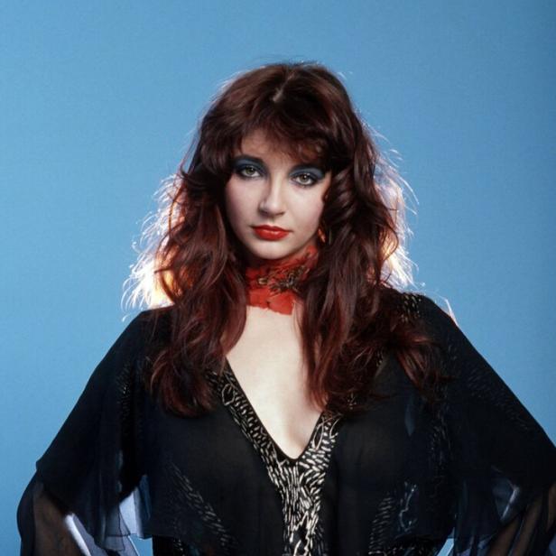 Kate Bush posiert vor einem blauen Hintergrund mit den Händen auf den Hüften.