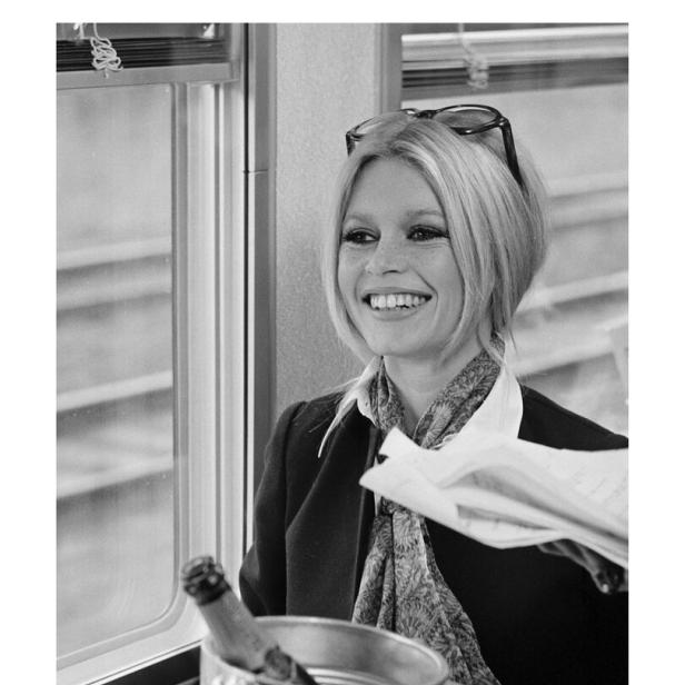 Brigitte Bardot lächelt in einem Zugabteil, mit einer Flasche Champagner im Vordergrund.