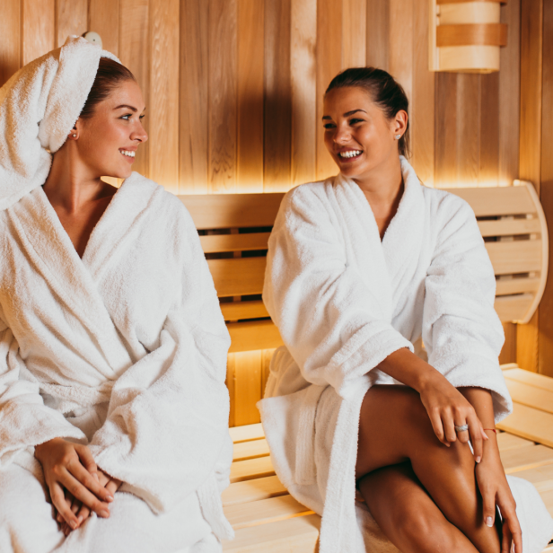 Zwei Frauen sitzen in Bademänteln in einer Sauna und lächeln sich an.