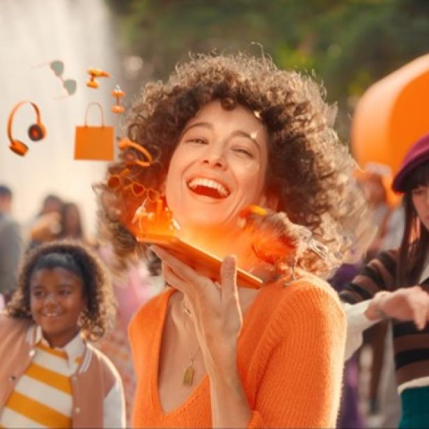 Eine Frau hält ein oranges Telefon in der Hand und lächelt freundlich in die Kamera.