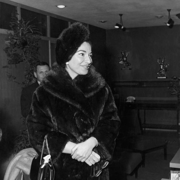 Maria Callas in einem Pelzmantel und einer Pelzmütze, die eine Handtasche und eine Brille hält.