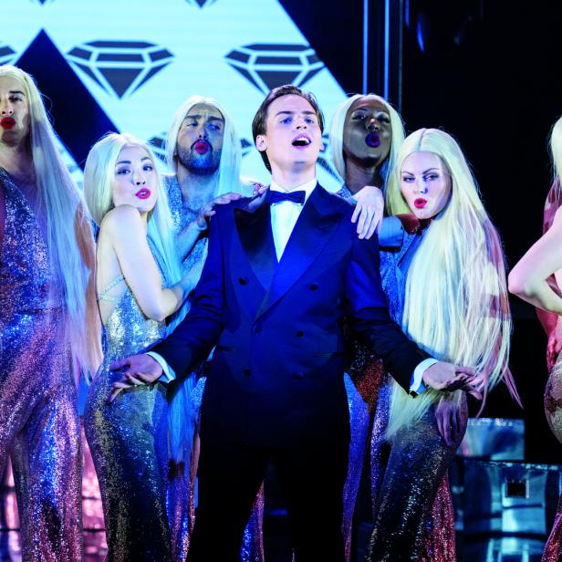 Ein Mann im Anzug singt, umgeben von Personen in glitzernden Kleidern und blonden Perücken.